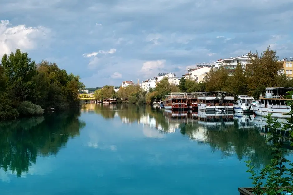 Peaceful riverside view with moored boats, lush greenery, and overcast sky in a serene town setting.